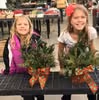 Two girls sitting at a picnic table smiling, each holding a decorated poinsettia plant with festive bows.