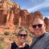 Smiling couple wearing sunglasses posing in front of red rock cliffs under a blue sky.