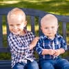 Two young boys sitting on a bench outdoors, both wearing plaid shirts and smiling.