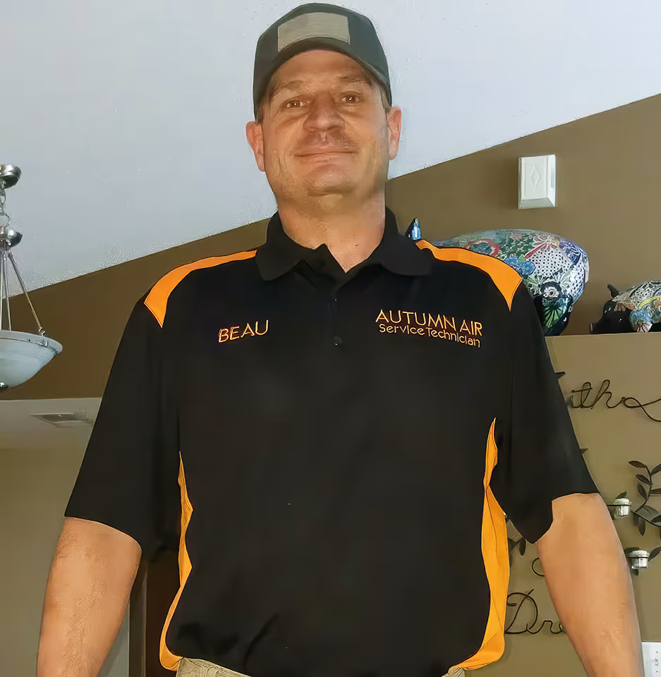 Man wearing a black and orange uniform shirt with 'BEAU' and 'AUTUMN AIR Service Technician' embroidered, and a baseball cap indoors.